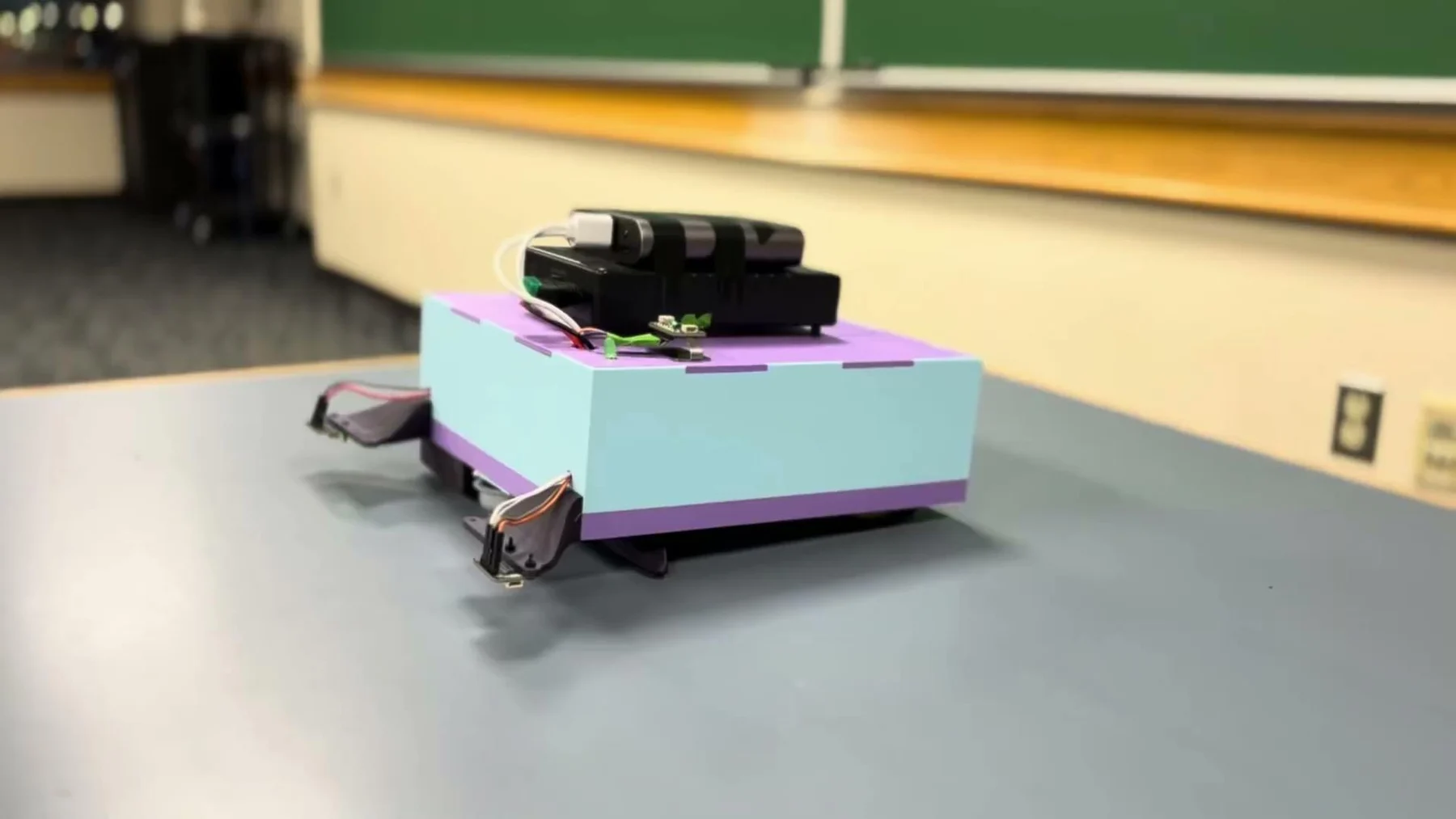 The assembled Deskinator robot on a tabletop beside its charging and test equipment.
